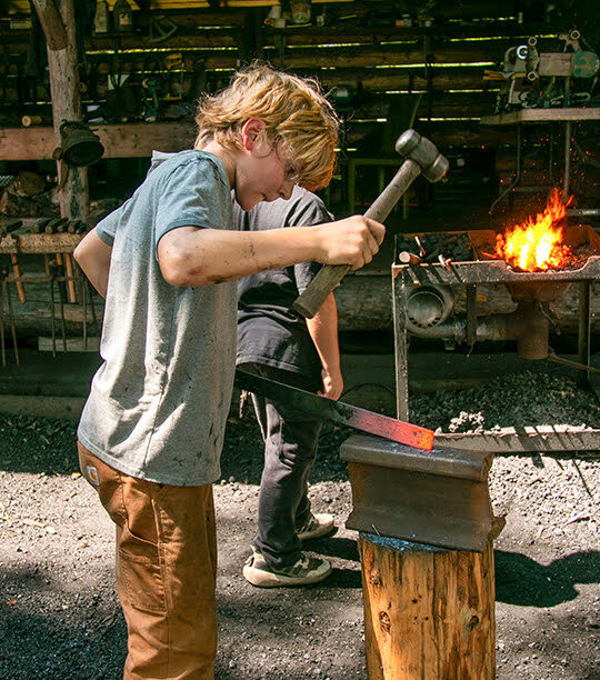 Campers learning blacksmithing techniques around outdoor anvil station
