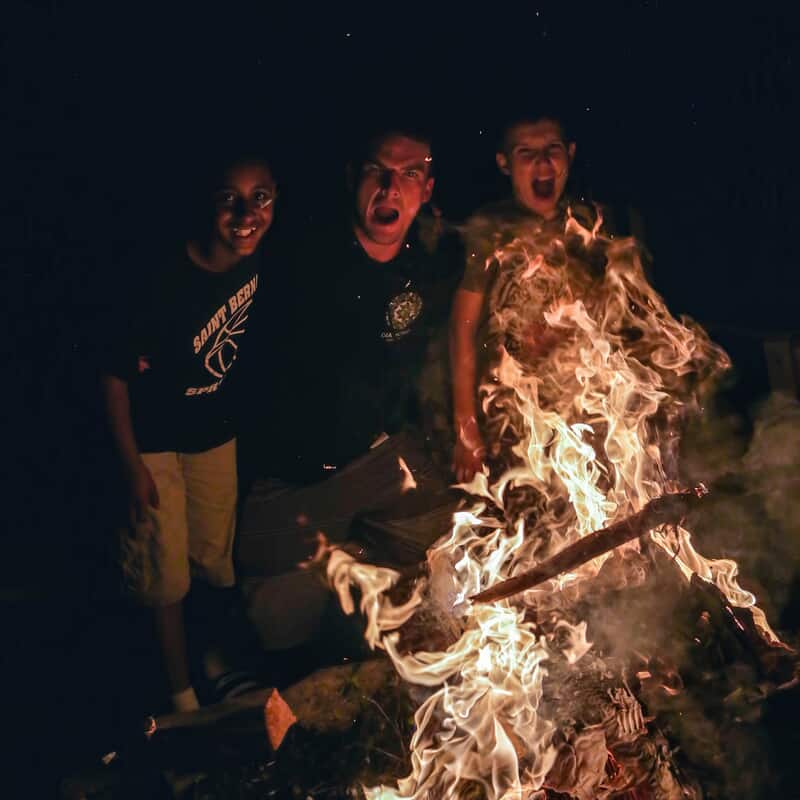 Campers gathered around a large nighttime campfire, smiling and reacting to the bright flames during an evening activity at summer camp.