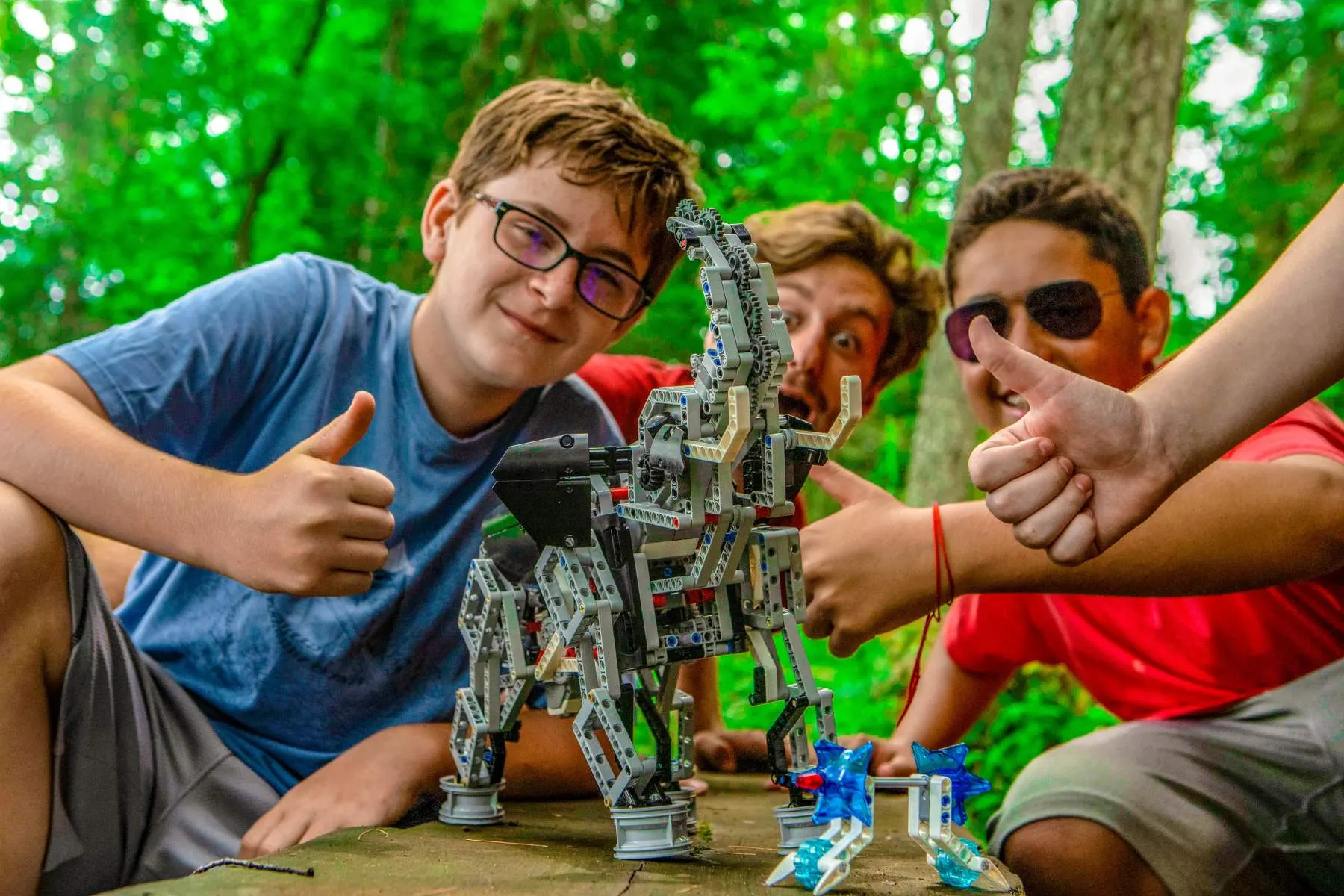 Campers proudly showing a large LEGO-style robot they built during an engineering activity, giving thumbs up in the forest workshop area.